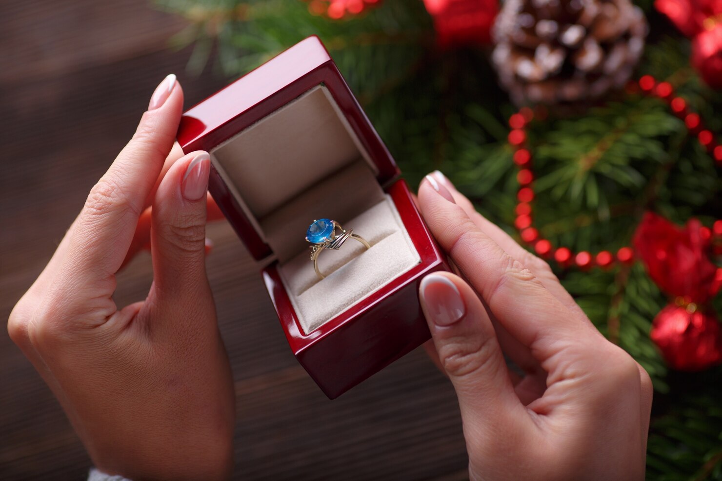 Hands holding a red jewelry box with a ring featuring a blue gemstone against a festive background.